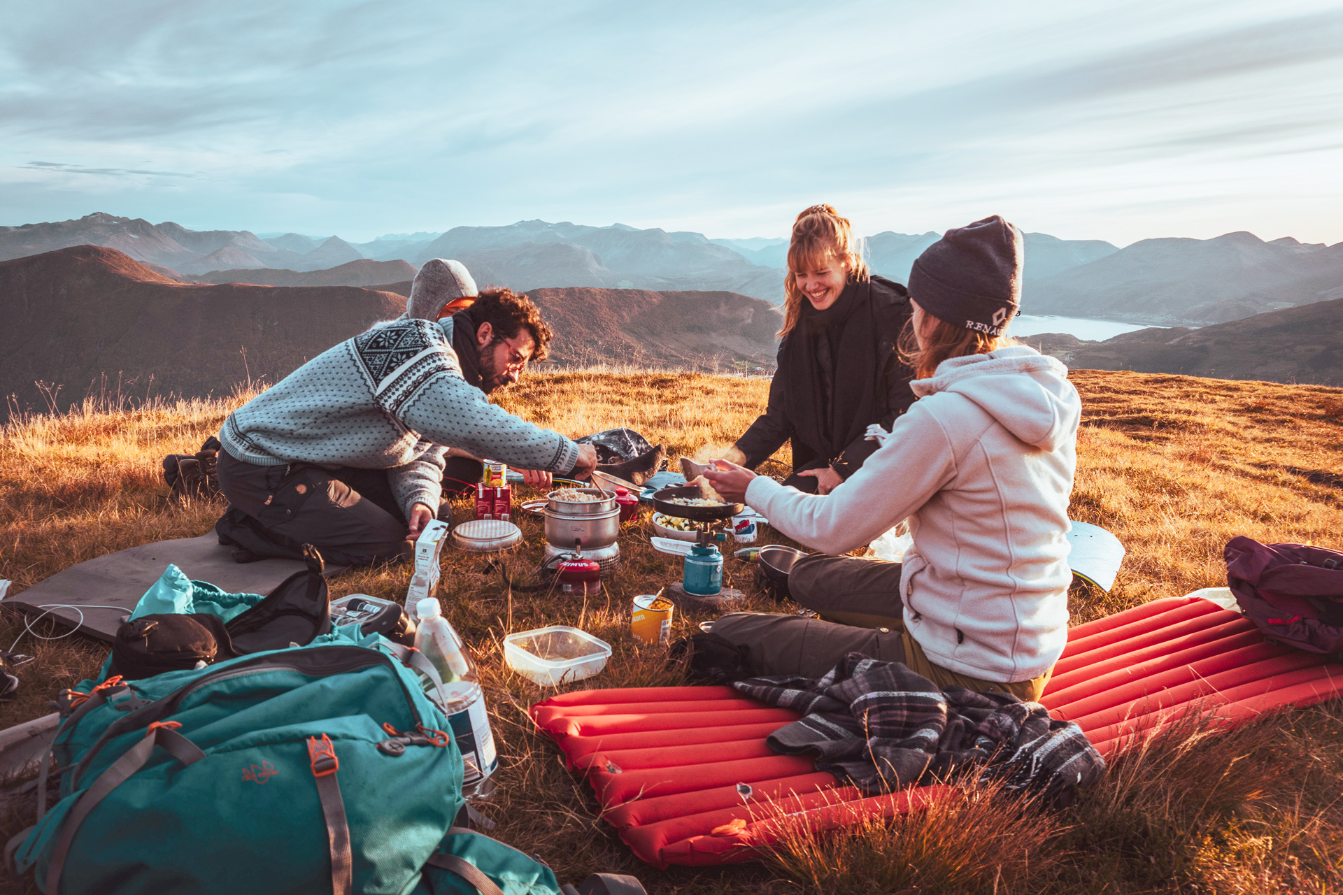 Junge Menschen sitzen mit Wanderausrüstung auf einem Bergplateau und bereiten Essen auf Campingkochern zu.
