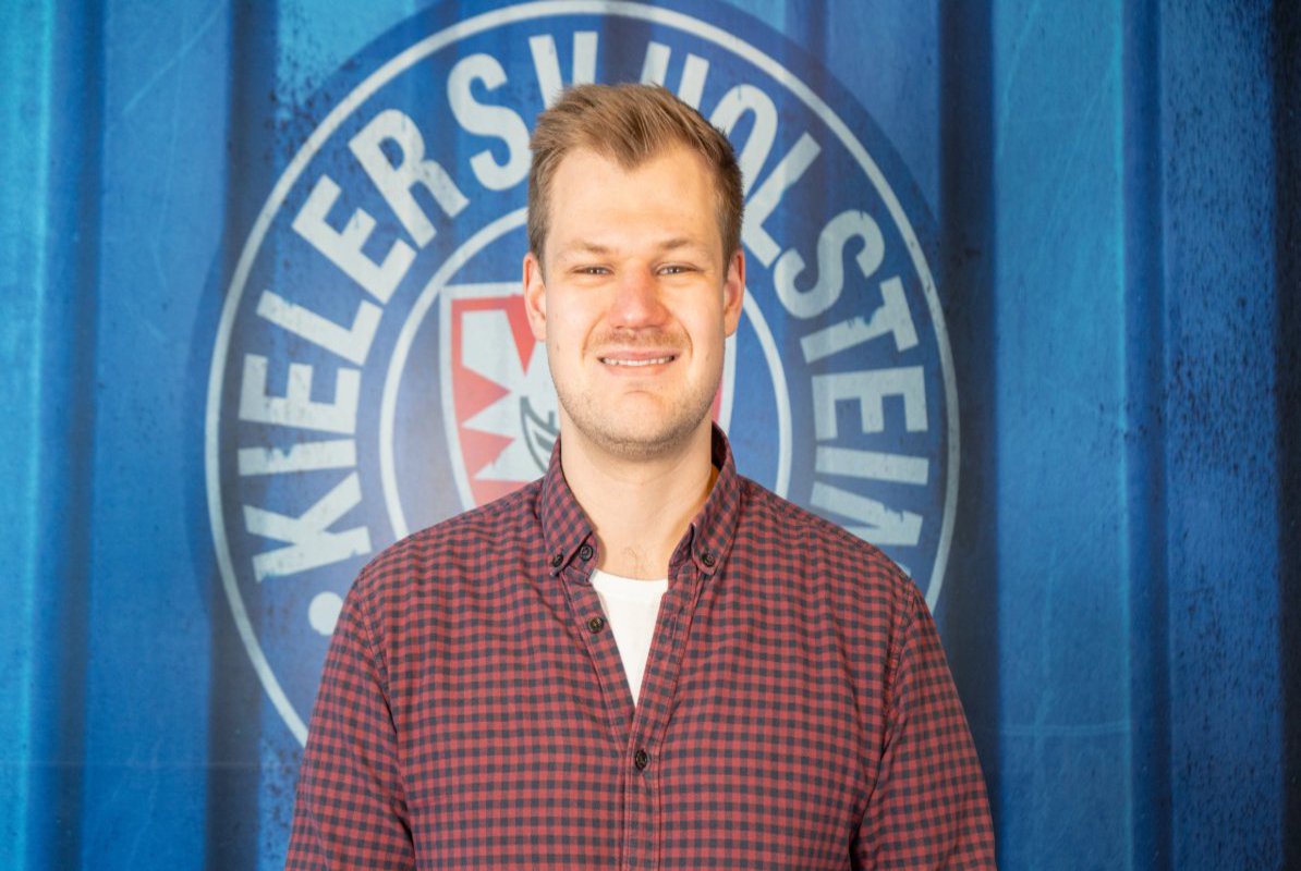 A man in a shirt poses in front of the KSV Holstein Kiel logo